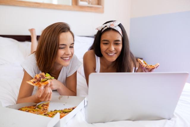 two women eating pizza in bed watching on a laptop